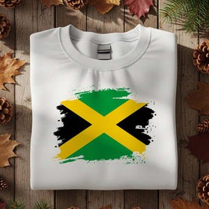 May include: White sweatshirt featuring the Jamaican flag design. The flag is rendered with a green background, a yellow cross, and black accents. The sweatshirt is folded and placed on a wooden surface with decorative elements.