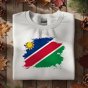 May include: White crew neck sweatshirt featuring a colorful graphic of the Namibian flag. The flag design includes blue, red, green, and yellow elements. The sweatshirt is folded on a wooden surface.