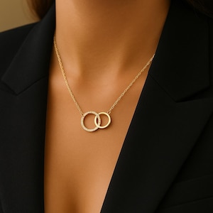 Op de afbeelding: Een goudkleurige ketting met twee in elkaar grijpende cirkels. Eén cirkel is versierd met kleine, heldere edelstenen, de andere is effen. De ketting wordt getoond tegen een donkere achtergrond, wat het ontwerp van de sieraden benadrukt.