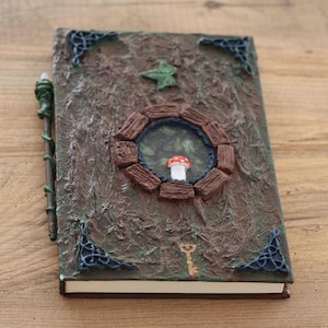 Può includere: Un diario a tema fantasy con una copertina marrone strutturata. Presenta una finestra circolare con un fungo in miniatura, una chiave e angoli decorativi. Una penna verde a forma di vite poggia sul lato. Il design del diario evoca un'estetica stravagante e boschiva.