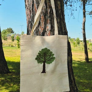 Op de afbeelding: Een beige canvas draagtas met een groen geborduurd boomontwerp. De tas heeft lange, crèmekleurige handvatten en hangt aan een boomstam. De achtergrond toont een park met bomen en een blauwe lucht.