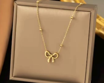 May include: A delicate gold-colored necklace featuring a bow pendant. The necklace has a fine chain with small, round accents. The bow pendant is centered and adds a touch of elegance. The necklace is displayed on a neutral-toned surface.