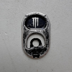 Może przedstawiać: Srebrno-czarna aluminiowa zawleczka od puszki z napojem, z białym logo Monster Energy nadrukowanym w górnej części. Metalowa powierzchnia wykazuje ślady użytkowania z widocznymi zarysowaniami i otarciami na wykończeniu oraz czarnych elementach.