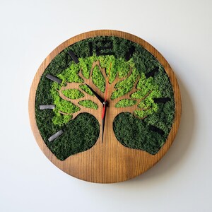 Könnte beinhalten: Eine runde Wanduhr aus Holz mit einem Baumdesign. Das Zifferblatt ist mit grünem Moos gefüllt, mit einer Baumsilhouette aus dem Holz geschnitten. Schwarze Zeiger und Stundenmarkierungen sind vor dem grünen Moos angeordnet. Die Uhr hat eine natürliche, rustikale Ästhetik.