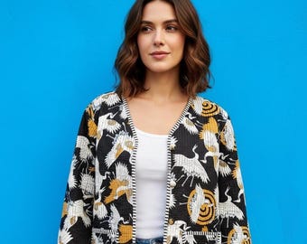 May include: A black jacket with a white and gold crane pattern. The jacket has a V-neck, two pockets, and striped cuffs. The model is wearing a white top and blue jeans.