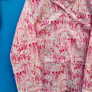 Może przedstawiać: R&oacute;żowy i biały zestaw piżamowy z powtarzającym się wzorem element&oacute;w architektonicznych i palm. Top z długim rękawem ma kołnierz, kieszeń na piersi i guziki. Tkanina wydaje się lekka.