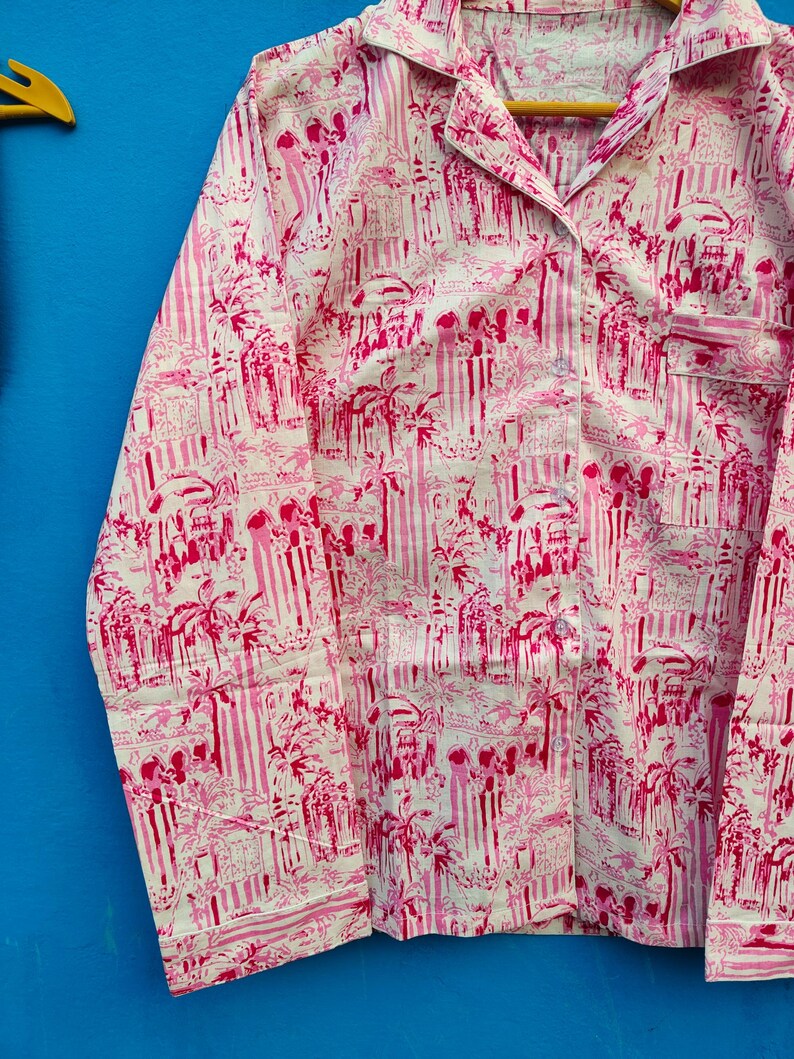 Może przedstawiać: R&oacute;żowy i biały zestaw piżamowy z powtarzającym się wzorem element&oacute;w architektonicznych i palm. Top z długim rękawem ma kołnierz, kieszeń na piersi i guziki. Tkanina wydaje się lekka.