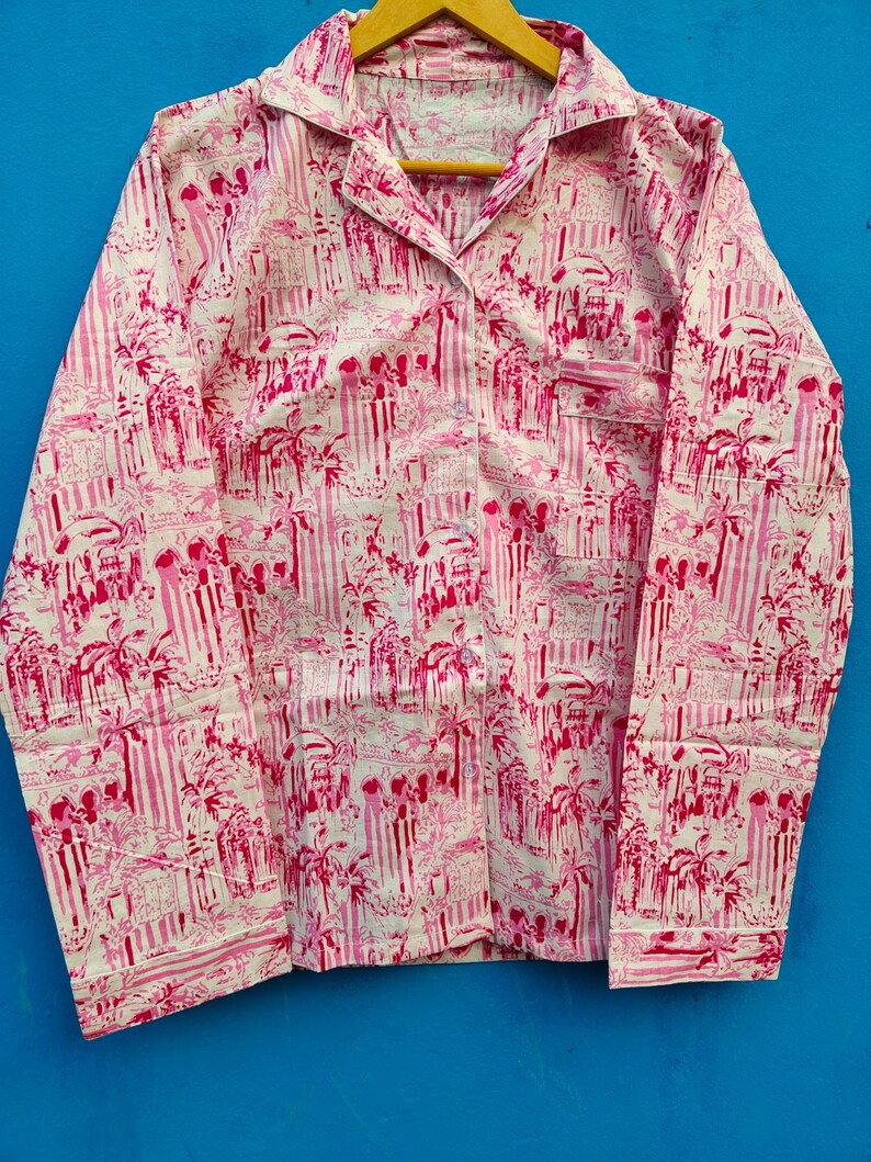 Może przedstawiać: R&oacute;żowa i biała koszula do piżamy z powtarzającym się wzorem pionowych pask&oacute;w oraz motyw&oacute;w architektonicznych i palm. Koszula z długim rękawem ma kołnierzyk, kieszeń na piersi i zapięcia na guziki.