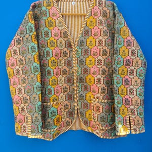 May include: A quilted jacket with a colorful floral pattern in shades of pink, yellow, blue, and gray. The jacket has a V-neck, two pockets, and a striped lining. The jacket is hanging on a wooden hanger.