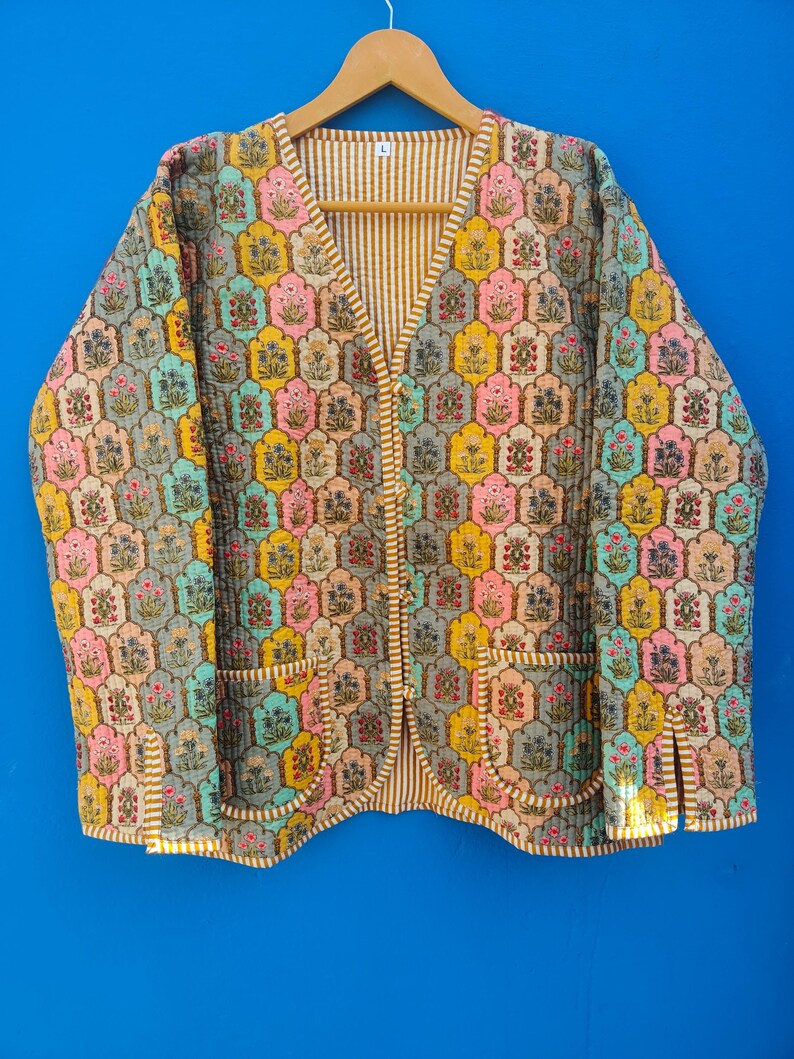 May include: A quilted jacket with a colorful floral pattern in shades of pink, yellow, blue, and gray. The jacket has a V-neck, two pockets, and a striped lining. The jacket is hanging on a wooden hanger.