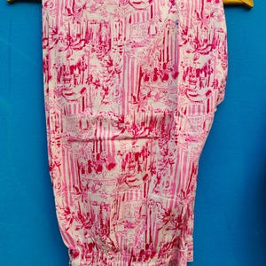 Może przedstawiać: R&oacute;żowe i białe spodnie od piżamy w wzory wiszące na ż&oacute;łtym drewnianym wieszaku na niebieskim tle. Spodnie mają powtarzający się wz&oacute;r pionowych pask&oacute;w oraz zdobnych element&oacute;w kwiatowych i architektonicznych.