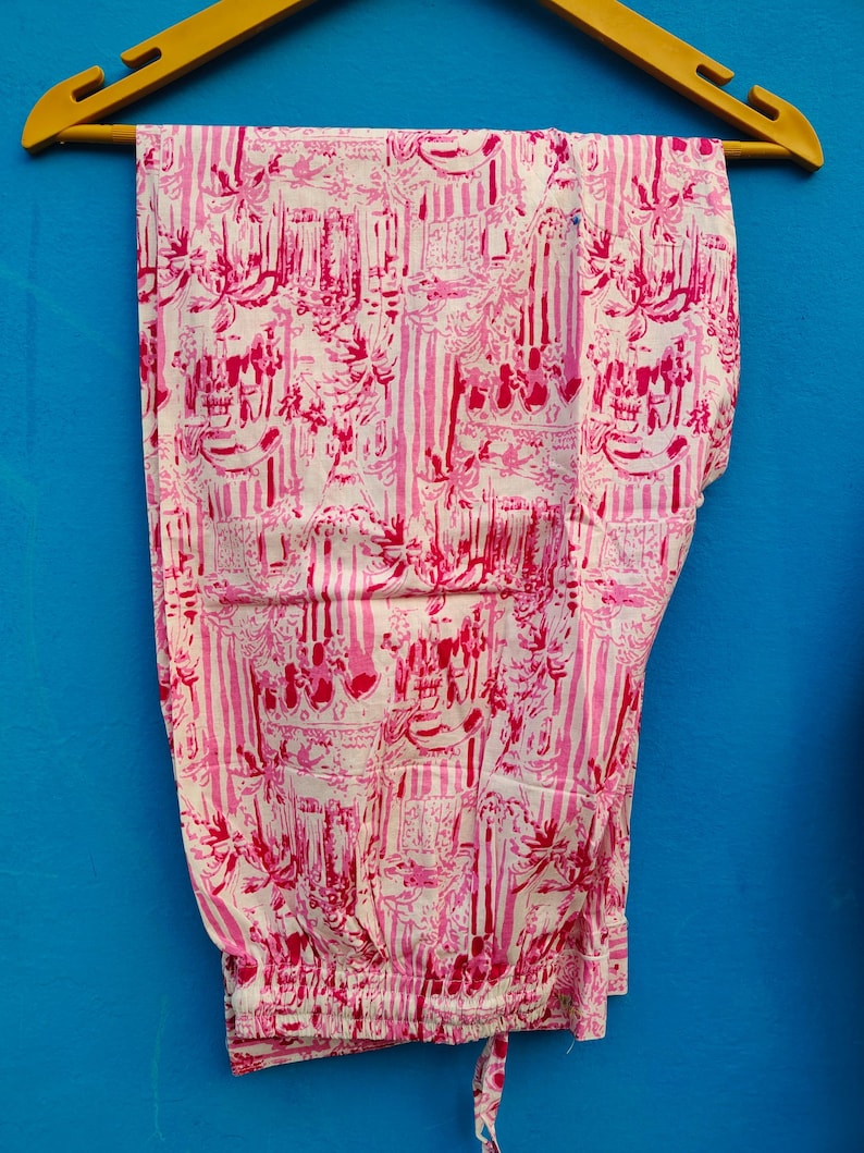 Może przedstawiać: R&oacute;żowe i białe spodnie od piżamy w wzory wiszące na ż&oacute;łtym drewnianym wieszaku na niebieskim tle. Spodnie mają powtarzający się wz&oacute;r pionowych pask&oacute;w oraz zdobnych element&oacute;w kwiatowych i architektonicznych.