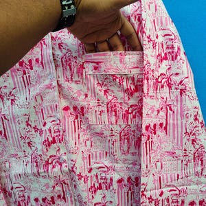 Może przedstawiać: R&oacute;żowa i biała koszula w tropikalny wz&oacute;r z palmami i elementami architektonicznymi. Koszula ma kieszeń na piersi i jest wykonana z lekkiego materiału. Koszula jest trzymana przez osobę.