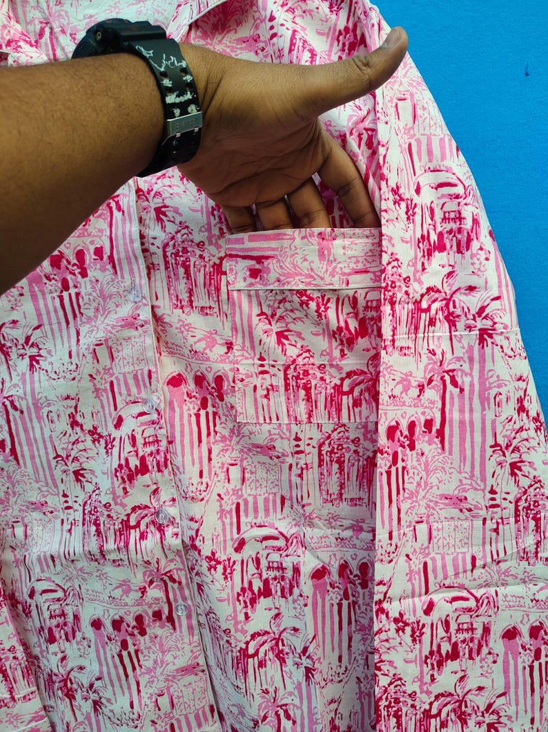 Może przedstawiać: R&oacute;żowa i biała koszula w tropikalny wz&oacute;r z palmami i elementami architektonicznymi. Koszula ma kieszeń na piersi i jest wykonana z lekkiego materiału. Koszula jest trzymana przez osobę.