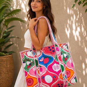 Puede incluir: Un bolso tote grande acolchado con un patrón vibrante y multicolor de frutas estilizadas, incluyendo granadas, limones y bayas. El bolso tiene una base blanca con detalles en rosa, azul, naranja y verde, acabado con un borde a rayas rosa y blanco.
