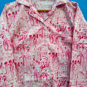 Może przedstawiać: R&oacute;żowa i biała koszula piżamowa z powtarzającym się wzorem budynk&oacute;w i palm. Koszula z długim rękawem ma kołnierzyk, kieszeń na piersi i guziki z przodu. Materiał wisi na drewnianym wieszaku.