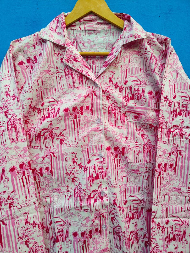 Może przedstawiać: R&oacute;żowa i biała koszula piżamowa z powtarzającym się wzorem budynk&oacute;w i palm. Koszula z długim rękawem ma kołnierzyk, kieszeń na piersi i guziki z przodu. Materiał wisi na drewnianym wieszaku.