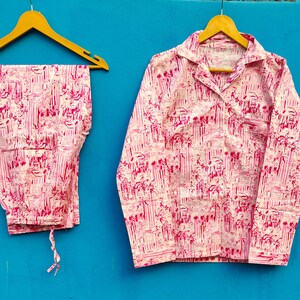 Może przedstawiać: R&oacute;żowy i biały zestaw piżamy z powtarzającym się wzorem budynk&oacute;w i drzew. Zestaw zawiera koszulę z długim rękawem zapinaną na guziki z kieszenią na piersi i pasujące spodnie. Piżama wisi na drewnianych wieszakach na niebieskim tle.