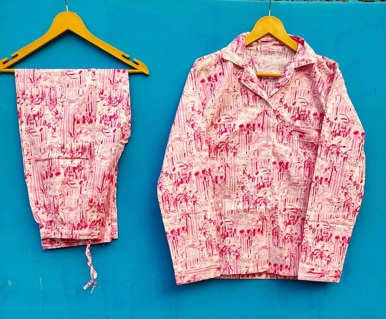 Może przedstawiać: R&oacute;żowy i biały zestaw piżamy z powtarzającym się wzorem budynk&oacute;w i drzew. Zestaw zawiera koszulę z długim rękawem zapinaną na guziki z kieszenią na piersi i pasujące spodnie. Piżama wisi na drewnianych wieszakach na niebieskim tle.