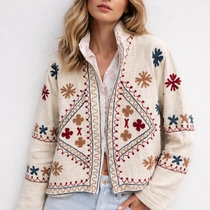 Pode incluir: Casaco bordado creme com gola alta. O casaco apresenta intrincados padrões florais e geométricos em fio vermelho, azul e castanho. O casaco é usado sobre uma camisa branca com gola e jeans azul claro.