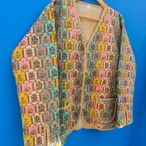 May include: A quilted jacket with a colorful floral pattern in shades of pink, yellow, blue, and gray. The jacket has a V-neck, two pockets, and a striped lining. It is hanging on a wooden hanger against a blue background.