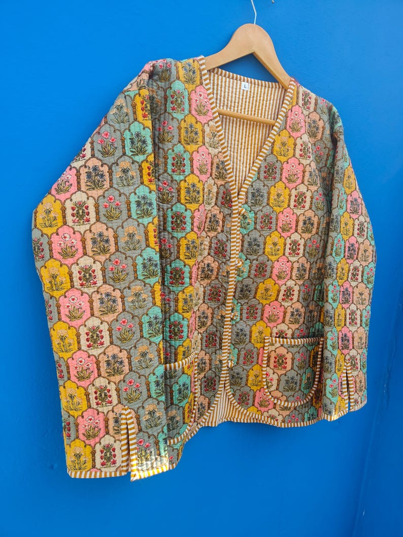 May include: A quilted jacket with a colorful floral pattern in shades of pink, yellow, blue, and gray. The jacket has a V-neck, two pockets, and a striped lining. It is hanging on a wooden hanger against a blue background.