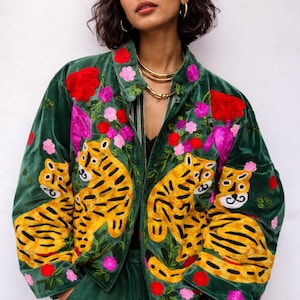 Pode incluir: Casaco de veludo verde com bordados de tigres e motivos florais. O casaco apresenta tigres amarelos com riscas pretas, flores vermelhas e rosa e um colarinho. A pessoa usa um colar dourado e calças verdes a condizer.