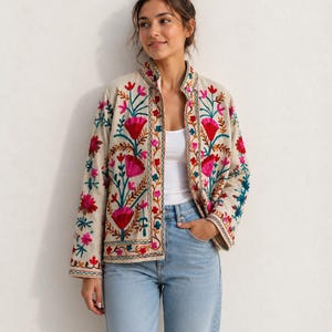 Könnte beinhalten: Eine beigefarbene Langarmjacke mit einem lebendigen Blumenstickmuster in Rosa-, Rot- und Petroltönen. Die Jacke hat einen Stehkragen, einen geraden Saum und eine durchgehende Knopfleiste, getragen über einem weißen Tanktop und einer hellblauen Jeans.