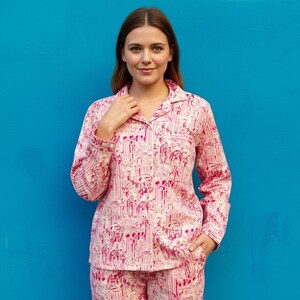 Może przedstawiać: R&oacute;żowy zestaw piżamowy z białym i r&oacute;żowym wzorem. Koszula z długim rękawem zapinana na guziki ma kołnierzyk i kieszeń na piersi. Widoczne są pasujące spodnie. Tło jest jednolicie niebieskie.