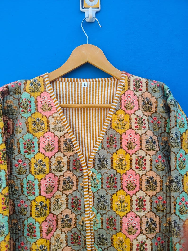 May include: A colorful, quilted jacket with a floral pattern in shades of yellow, pink, blue, and gray. The jacket has a V-neck and is lined with a striped fabric. The size "L" is visible on the hanger.