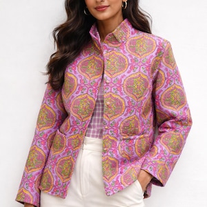 Puede incluir: Chaqueta rosa con estampado de cachemira en amarillo, verde y rosa. La chaqueta tiene cuello mandarín, bolsillos y botones dorados. La modelo lleva pantalones blancos y una camisa a cuadros debajo.