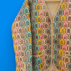 May include: A quilted jacket with a colorful floral pattern. The jacket features a V-neck, two pockets, and a striped lining. The floral design includes pink, yellow, blue, and peach tones.