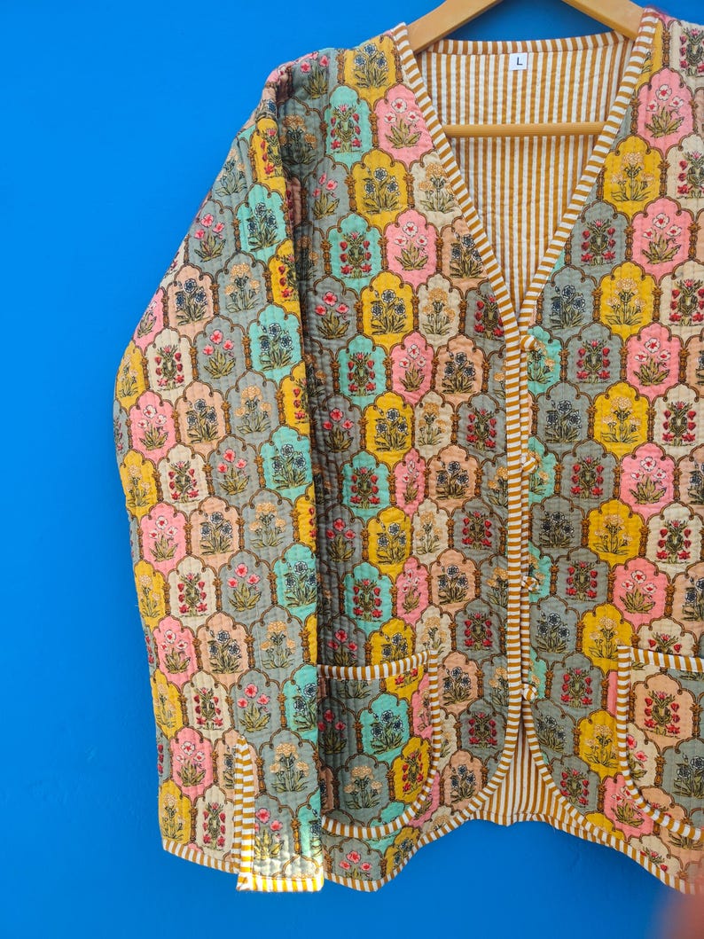 May include: A quilted jacket with a colorful floral pattern. The jacket features a V-neck, two pockets, and a striped lining. The floral design includes pink, yellow, blue, and peach tones.