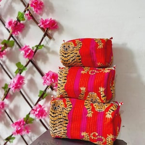 Può includere: Una pila di tre pochette rettangolari in tessuto di varie dimensioni, caratterizzate da un vivace motivo a righe rosse e rosa con una stampa tigre ripetuta. Ogni pochette presenta una consistenza trapuntata e piccole linguette in tessuto bianco agli angoli.