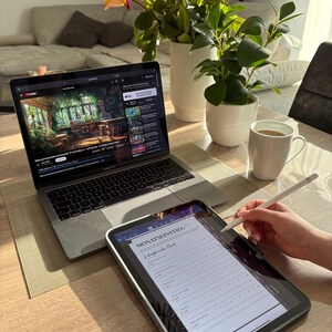 Pu&ograve; includere: Un laptop con un video di YouTube e un tablet con la scritta "MONATSEINSTIEG" sono su un tavolo di legno. Una tazza bianca con caff&egrave;, un vaso di fiori e una piccola pianta in vaso sono anch'essi presenti. Una mano usa uno stilo sul tablet.
