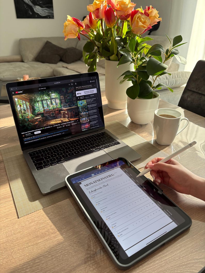 Pu&ograve; includere: Un laptop con un video di YouTube e un tablet con la scritta "MONATSEINSTIEG" sono su un tavolo di legno. Una tazza bianca con caff&egrave;, un vaso di fiori e una piccola pianta in vaso sono anch'essi presenti. Una mano usa uno stilo sul tablet.