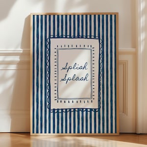 Puede incluir: Impresión artística enmarcada con un fondo a rayas azules y blancas. La obra presenta un rectángulo blanco con las palabras "Splish Splash" en escritura cursiva, enmarcado por un borde festoneado. El marco es de color madera clara.