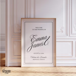 May include: A framed wedding welcome sign with the names "Emma & James" in elegant script. The sign includes the wedding date and location, "Château des Lavandes, Provence, France." The frame is gold, and the background is white.