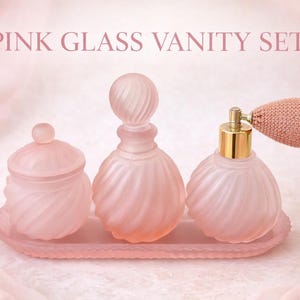 May include: Pink glass vanity set with a jar, perfume bottle, and atomizer on a tray. The set has a swirled design and is in a soft pink color. The text "PINK GLASS VANITY SET" is displayed at the top of the image.
