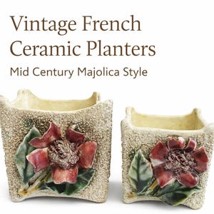 May include: Two vintage French ceramic planters in a Mid Century Majolica style. The planters are square with a textured, speckled exterior and a cream-colored interior. Each planter features a detailed floral design with red and green accents. The text reads "Vintage French Ceramic Planters".