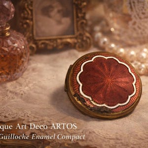 Puede incluir: Un compacto Art Déco antiguo con un diseño de esmalte guilloché rojo y un borde festoneado blanco. El compacto es redondo y descansa sobre una superficie de encaje. Un frasco de perfume y un marco dorado están en el fondo.