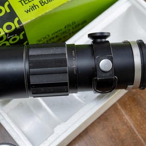 Puede incluir: Un objetivo teleobjetivo Soligor negro de 300 mm f/5.6 con parasol incorporado. La lente está en una caja blanca y tiene un anillo plateado y una correa negra. La caja es verde y blanca.