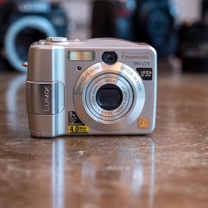 May include: Silver Panasonic DMC-LC70 digital camera with a Leica lens. The camera has a 3x optical zoom and 4.0 megapixels. The camera is on a brown surface with other cameras in the background.