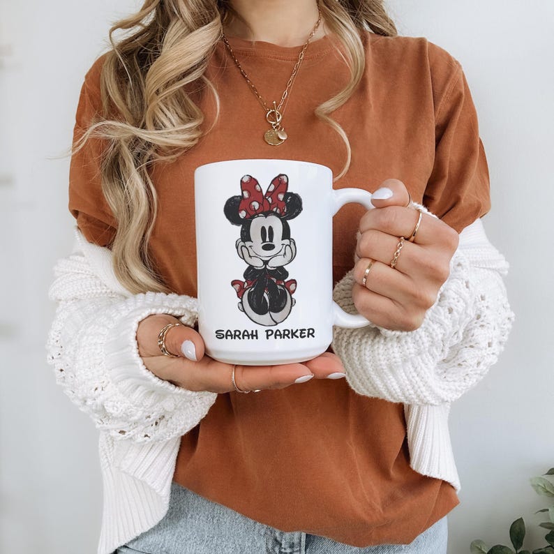 May include: White ceramic mug featuring a Minnie Mouse design with a red bow and black details. The mug has the name "SARAH PARKER" printed in black. The mug is held by a person wearing a rust-colored shirt and a white cardigan.
