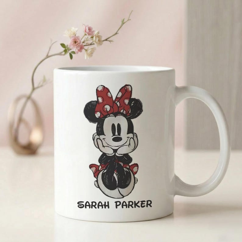May include: White ceramic mug featuring a black and red Minnie Mouse design. Minnie Mouse is depicted with a red bow with white polka dots. The name "SARAH PARKER" is printed in black at the bottom of the mug.