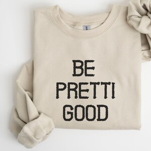 May include: A beige sweatshirt with the phrase "BE PRETTI GOOD" printed in black block letters. A matching beige scrunchie rests beside the folded sleeve. The sweatshirt is made of a soft material.