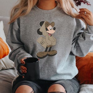 May include: A gray sweatshirt featuring a cartoon Mickey Mouse character in a brown hoodie and shoes, holding a shopping bag with the Gucci logo. The sweatshirt is a solid gray color.