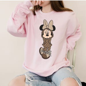 May include: A light pink sweatshirt featuring a Minnie Mouse graphic. The cartoon character is dressed in a brown outfit with a repeating pattern and matching bow. The sweatshirt has long sleeves and a crew neck.