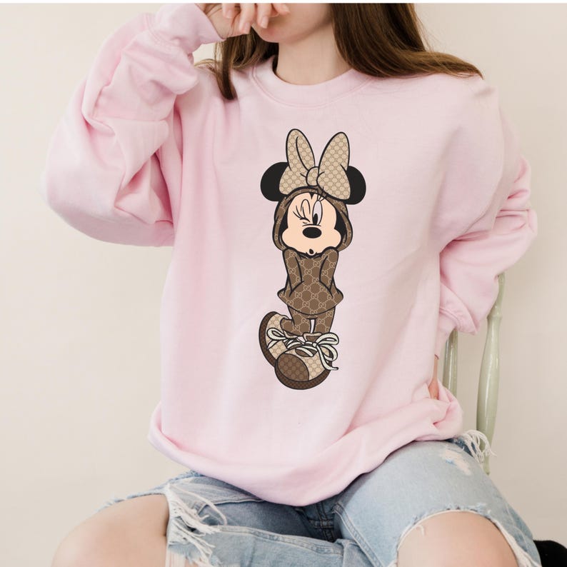 May include: A light pink sweatshirt featuring a Minnie Mouse graphic. The cartoon character is dressed in a brown outfit with a repeating pattern and matching bow. The sweatshirt has long sleeves and a crew neck.