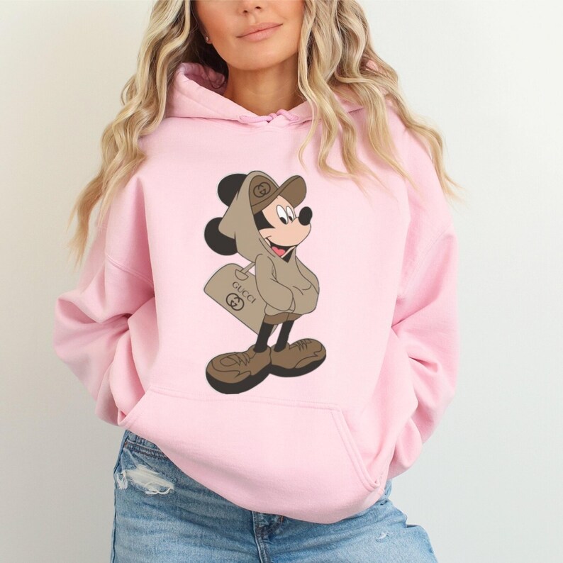May include: A light pink hoodie featuring a cartoon Mickey Mouse character wearing a brown hat and hoodie, holding a shopping bag with the Gucci logo. The character is in brown shoes. The hoodie has a front pocket.